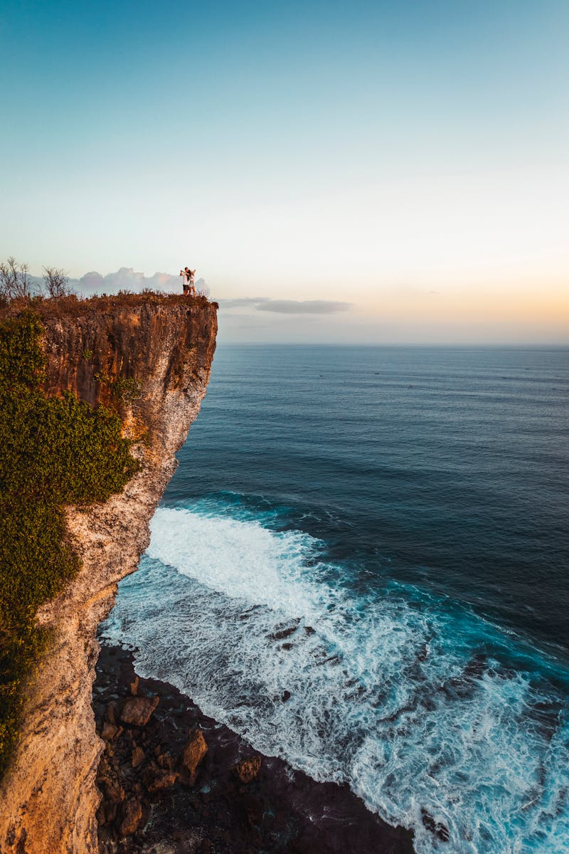 A couple stands on a dramatic cliff edge overlooking the ocean at sunset in Bali, Indonesia.