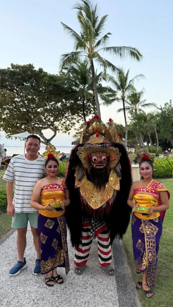 Tourist posing with Barong dance performers in traditional Balinese costumes amid palm trees.